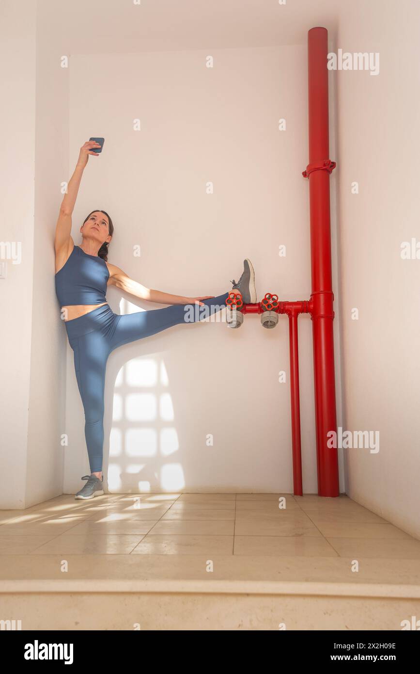 Mujer deportiva tomando un selfie en su teléfono mientras hace un estiramiento de la pierna contra un hidrante de fuego rojo Foto de stock