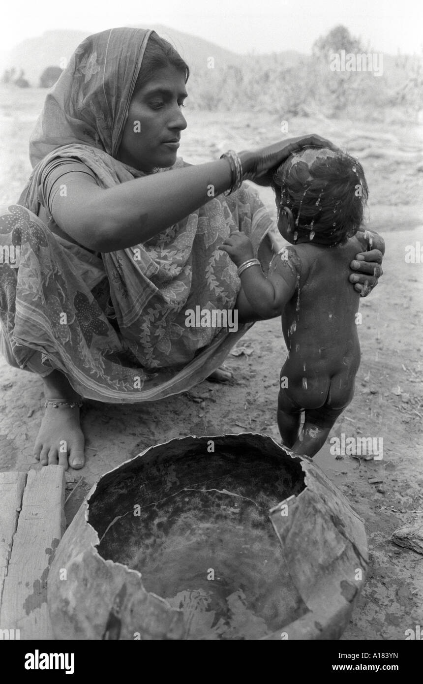 B/W de una trabajadora agrícola rural que lava a su bebé de una olla de agua. Gujarat, India Foto de stock