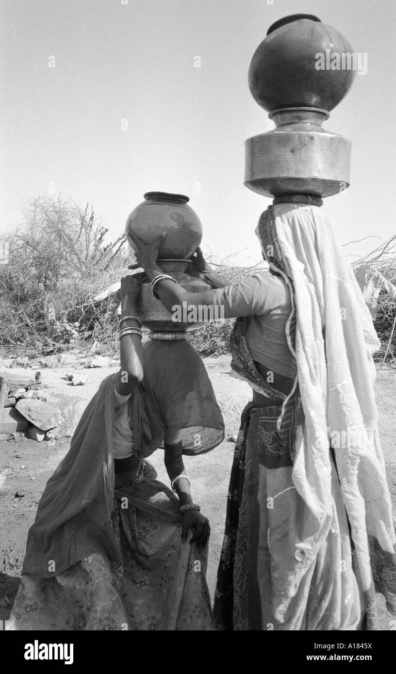 B/W de dos mujeres cargando ollas llenas de metal y agua de arcilla en sus cabezas después de llenar en el pozo, una tarea diaria para las mujeres rurales en Gujarat, India Foto de stock