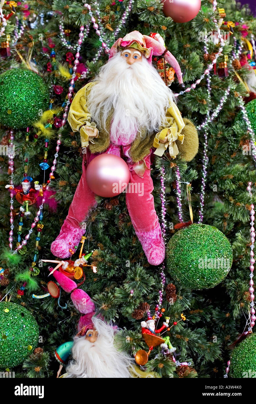 Santa colgando de un árbol de navidad decorado colorido con un baubel entre sus piernas y un sorprendido mira en su rostro. Foto de stock
