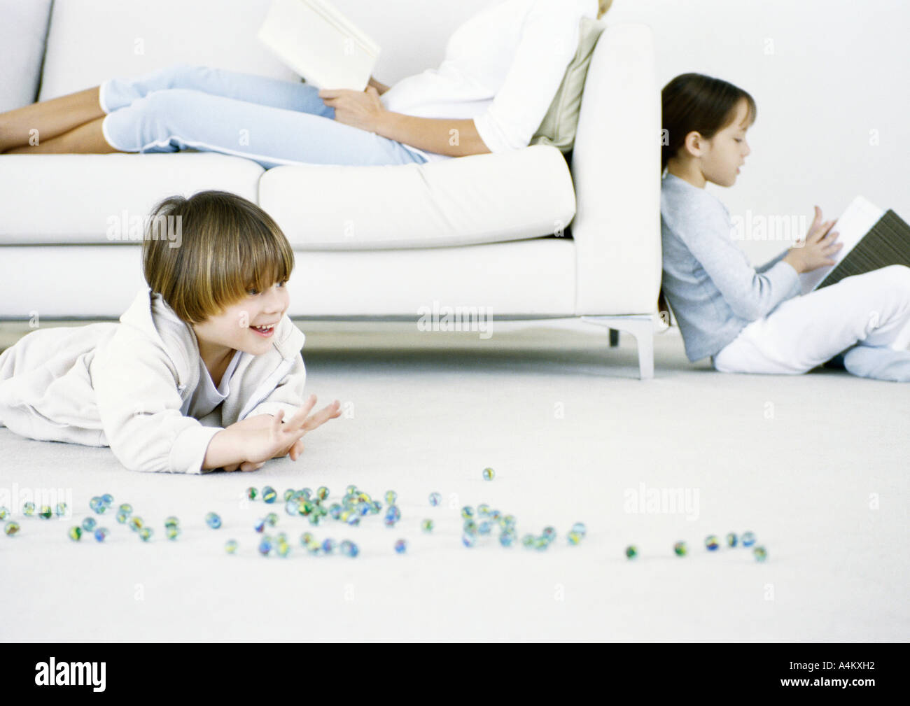 Niño jugando canicas en el suelo, la mujer y la niña leyendo en segundo plano. Foto de stock