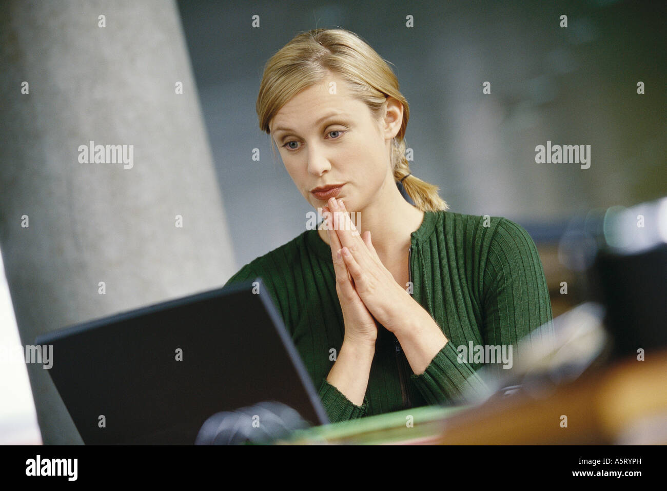 Mujer joven con laptop, las manos entrelazadas delante de barbilla Foto de stock