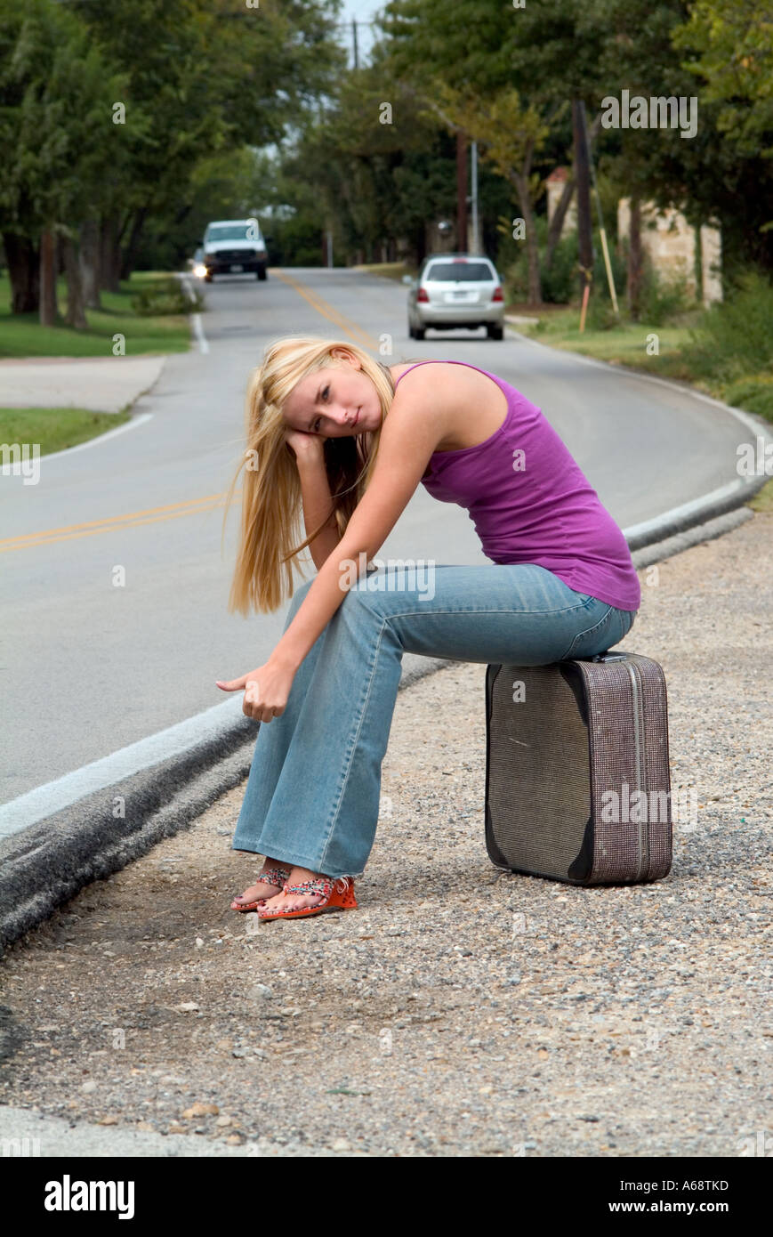 Joven chica rubia con pelo largo de los vaqueros sentados en una maleta al  lado de la carretera en busca de una forma de salir de la ciudad Fotografía  de stock -