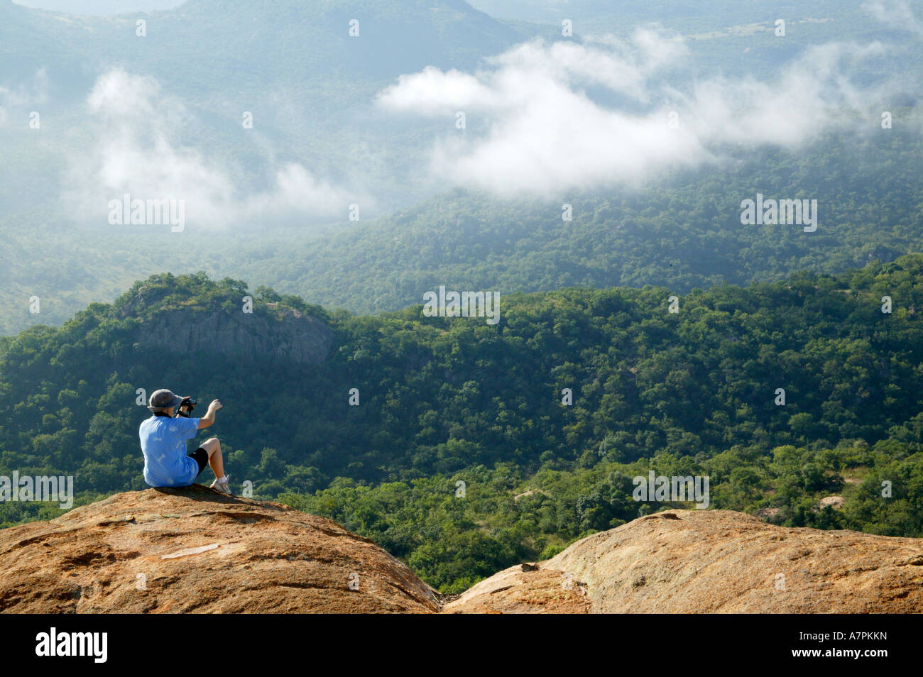 Una persona sentada en el borde de un afloramiento de granito apuntando hacia abajo el valle cubierto de vegetación autóctona Foto de stock