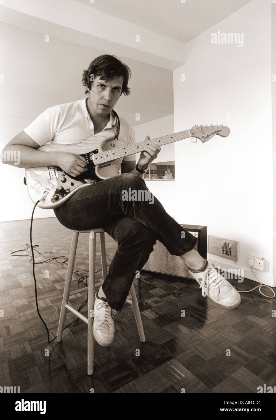 En blanco y negro de un hombre tocando una guitarra Fender Stratocaster sentado en un taburete con las piernas cruzadas en una habitación vacía Vibrolux amp Foto de stock