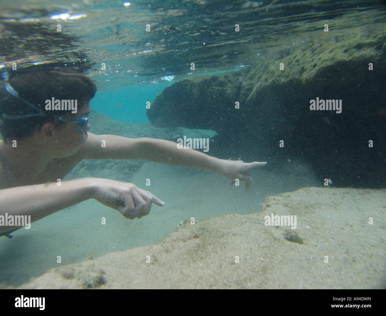 Fotografía submarina de un muchacho con gafas de natación y buceo apuntando debajo de una roca en el agua poco profunda Foto de stock