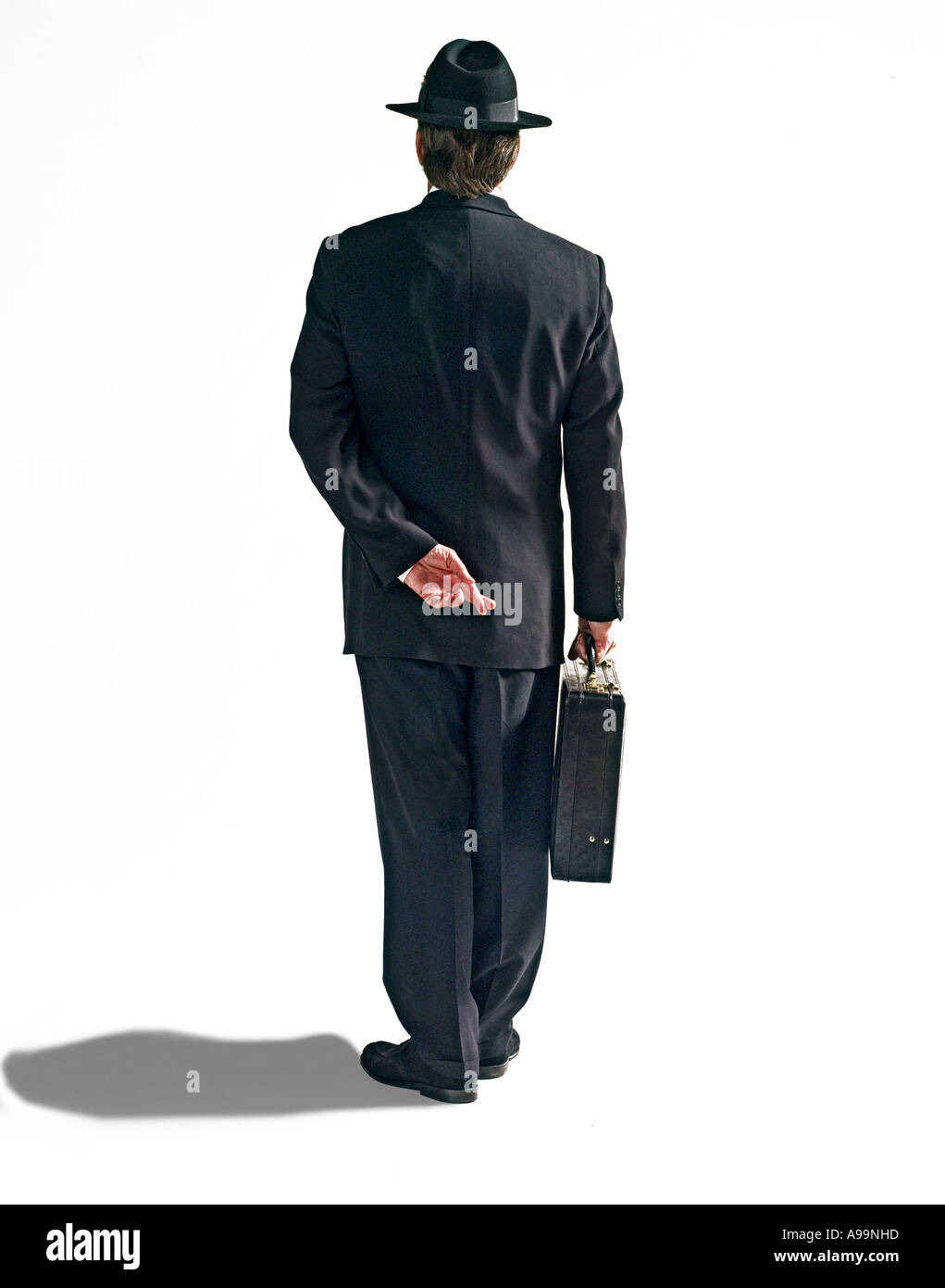 Hombre de negocios en traje negro con sombrero Fedora negro y el maletín con los dedos cruzados detrás de su espalda Foto de stock