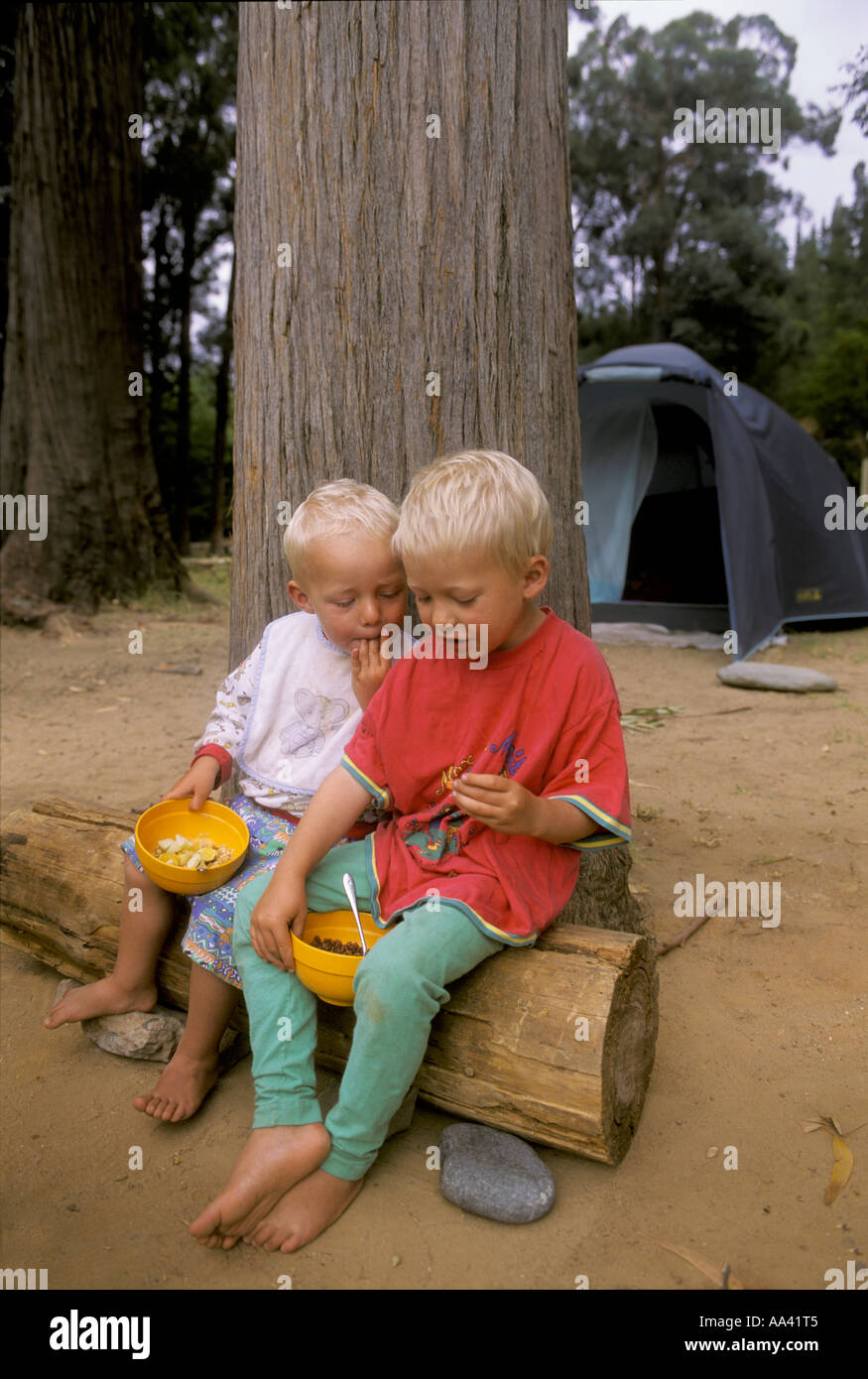 Niños pequeños, de dos y cuatro años, están sentados en un tronco para comer muesli, camping en Sudáfrica Foto de stock
