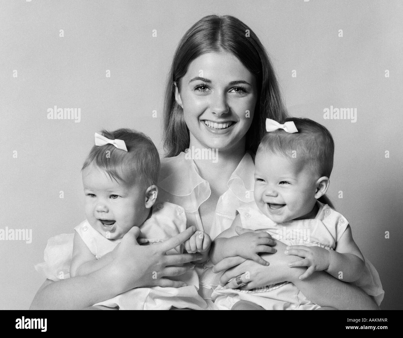 1970 Madre sonriente Celebración gemelos HEMBRA CON ARCOS EN CABELLO CON CADA BRAZO mirando a la cámara Foto de stock