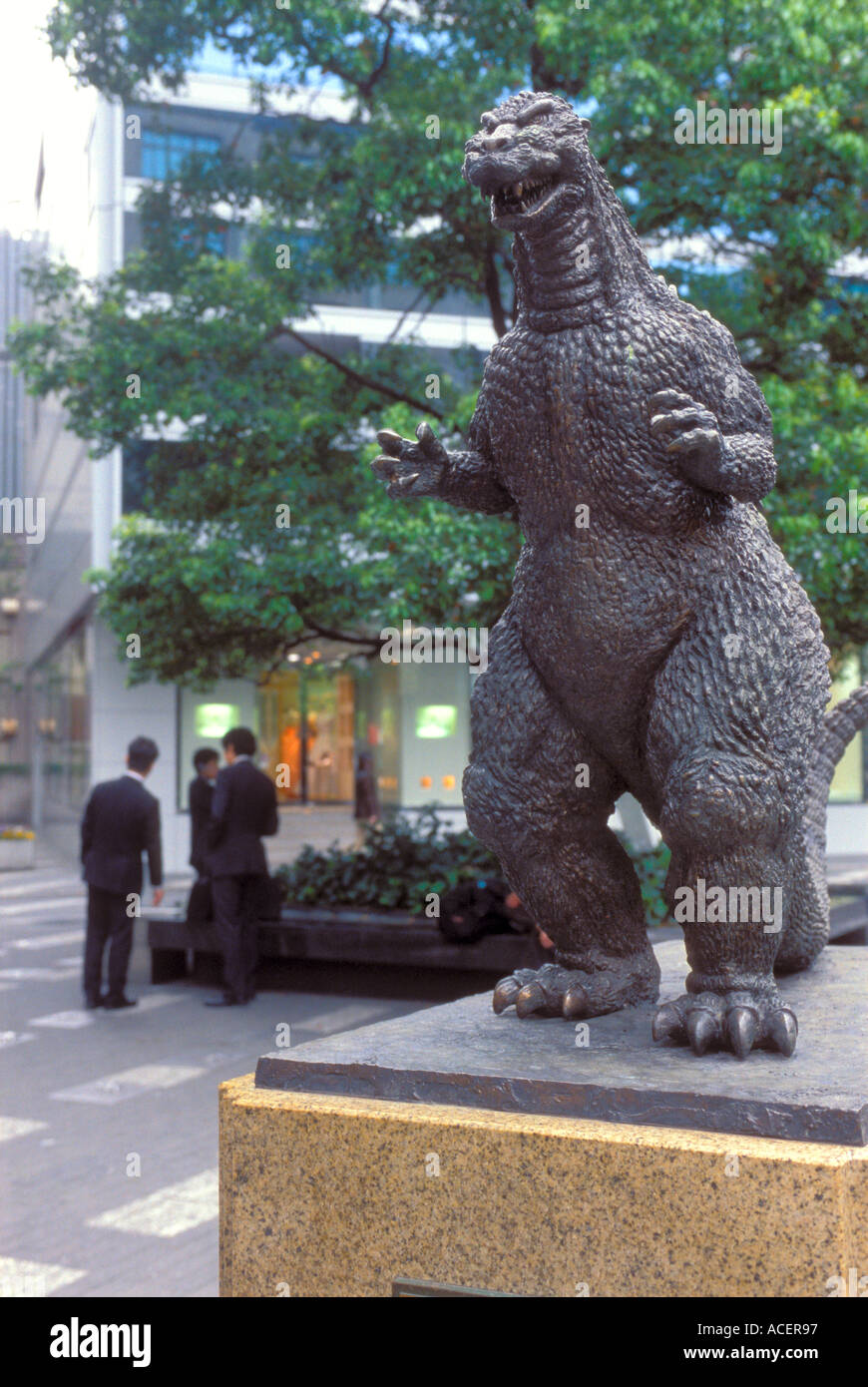 Estatua de bronce de Godzilla mirando a dos hombres de negocios en un parque tomando un descanso en el distrito de Yurakucho de Tokyo. Foto de stock