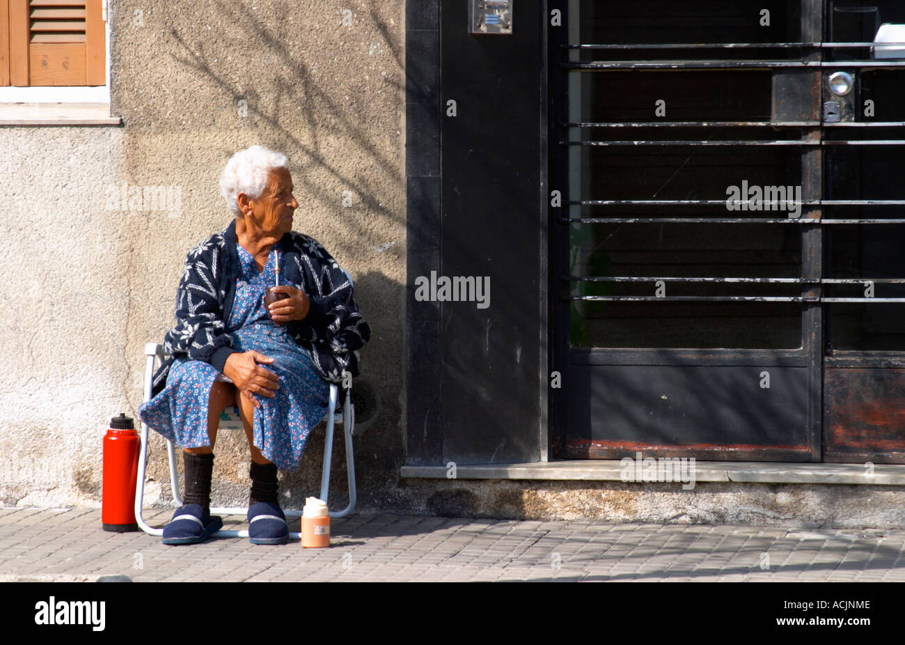 Una anciana sentada en la acera bebiendo mate té de hierbas, con un frasco  de agua caliente termo rojo delante de una puerta Puerta de hierro negro.  Montevideo, Uruguay, América del Sur