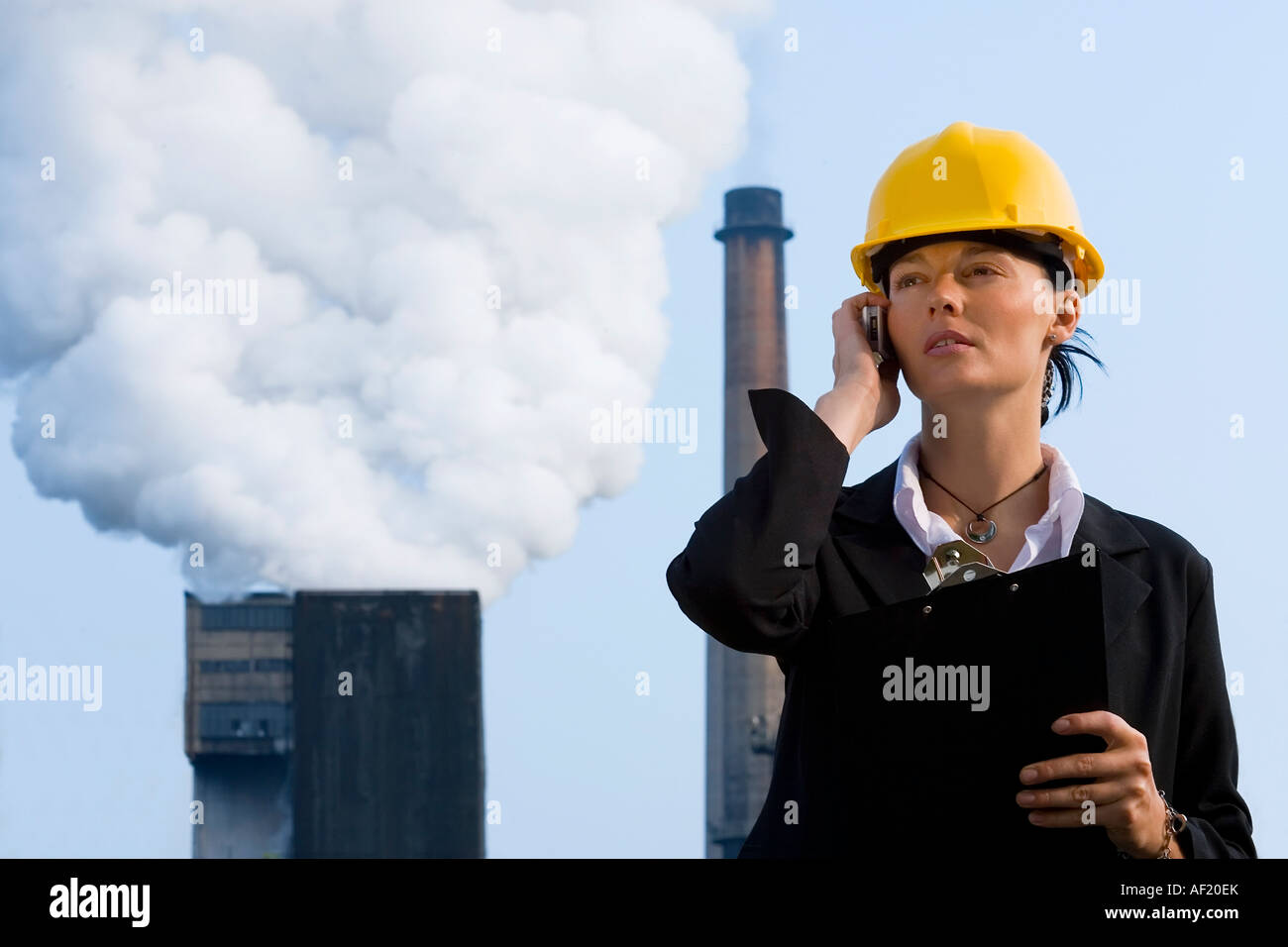 Una hermosa mujer de pelo oscuro en su teléfono móvil de pie delante de una fábrica de bombear hacia afuera la contaminación de sus chimeneas Foto de stock
