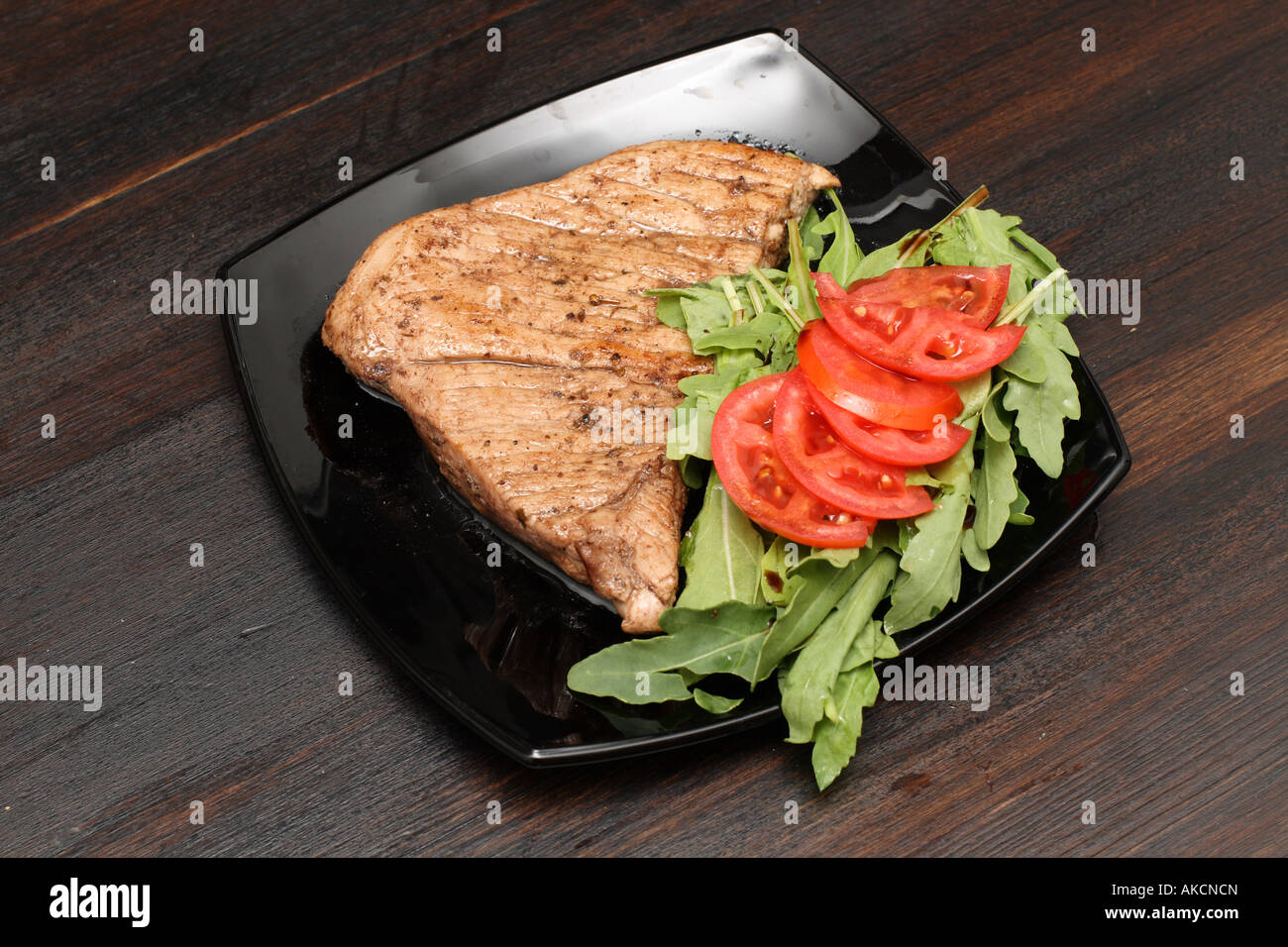 Porterhouse steak con tomates y verdes en la placa negra sobre mesa de madera oscura Foto de stock