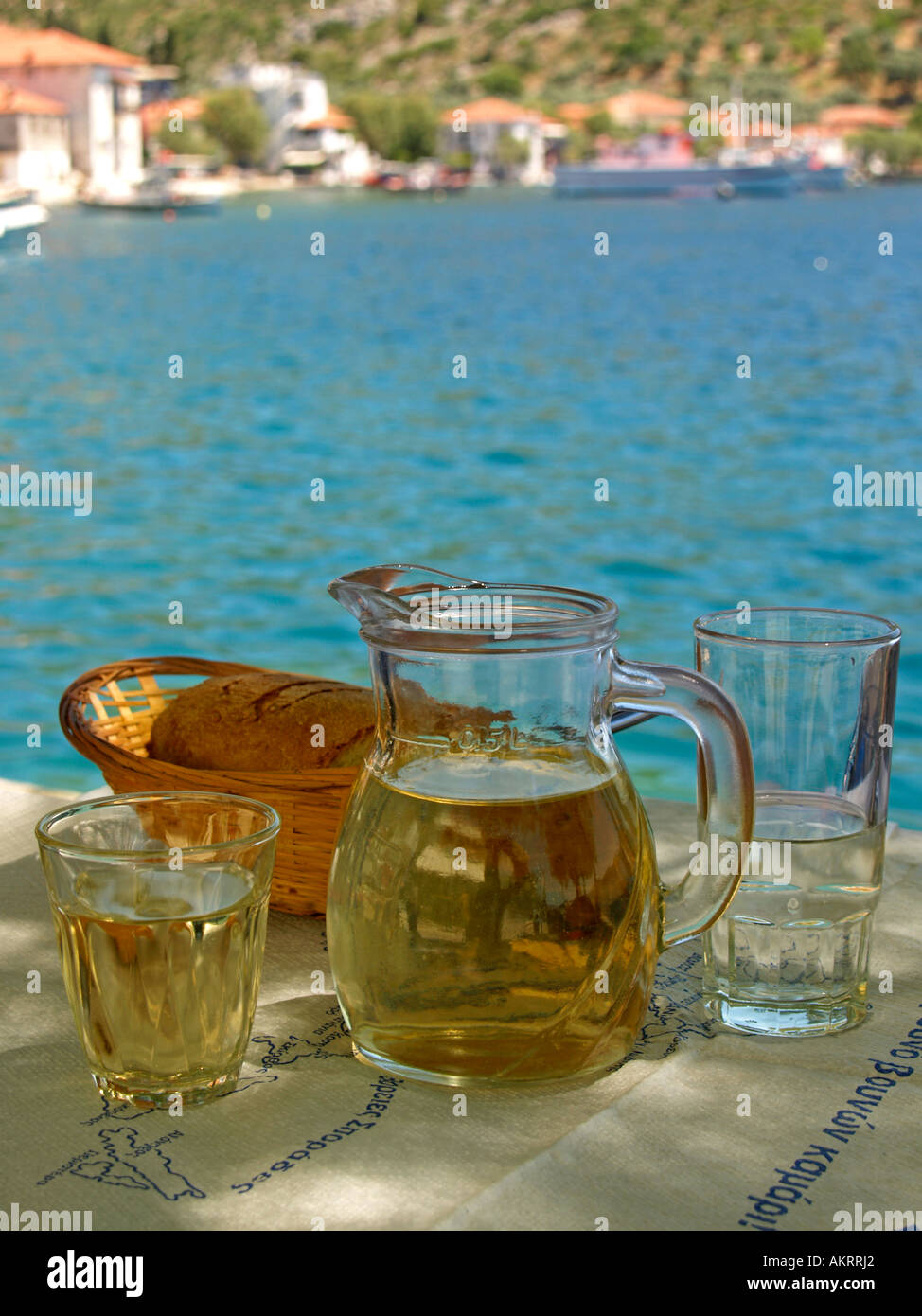 Una jarra llena de vino blanco Retsina dos vasos cesta con pan sobre una tabla en el fondo color turquesa del Mar Mediterráneo Foto de stock