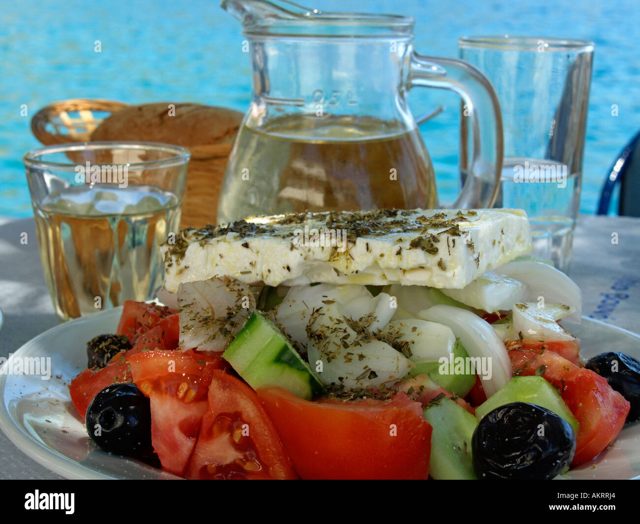 Ensalada griega una jarra llena de vino blanco Retsina cesta con pan sobre una tabla en el fondo color turquesa del Mar Mediterráneo Foto de stock
