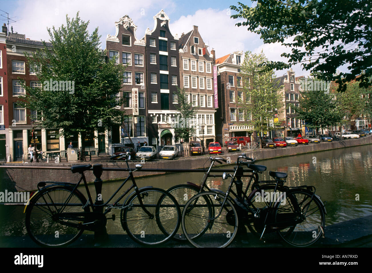 Negro bicicletas en Amsterdam inclinada contra las barandas de un puente con una línea de automóviles estacionados a lo largo de la arbolada canalside allá en frente del Museo Amstelkring originalmente una iglesia clandestina de los siglos XVII y XVIII, Foto de stock