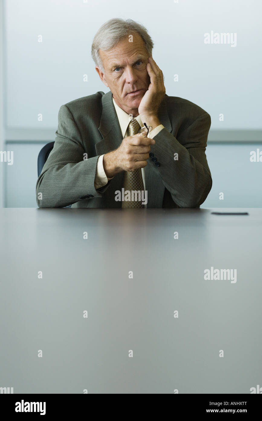 Empresario sentado con la mano bajo el mentón, sujetando las gafas en la mano, mirando a la cámara Foto de stock