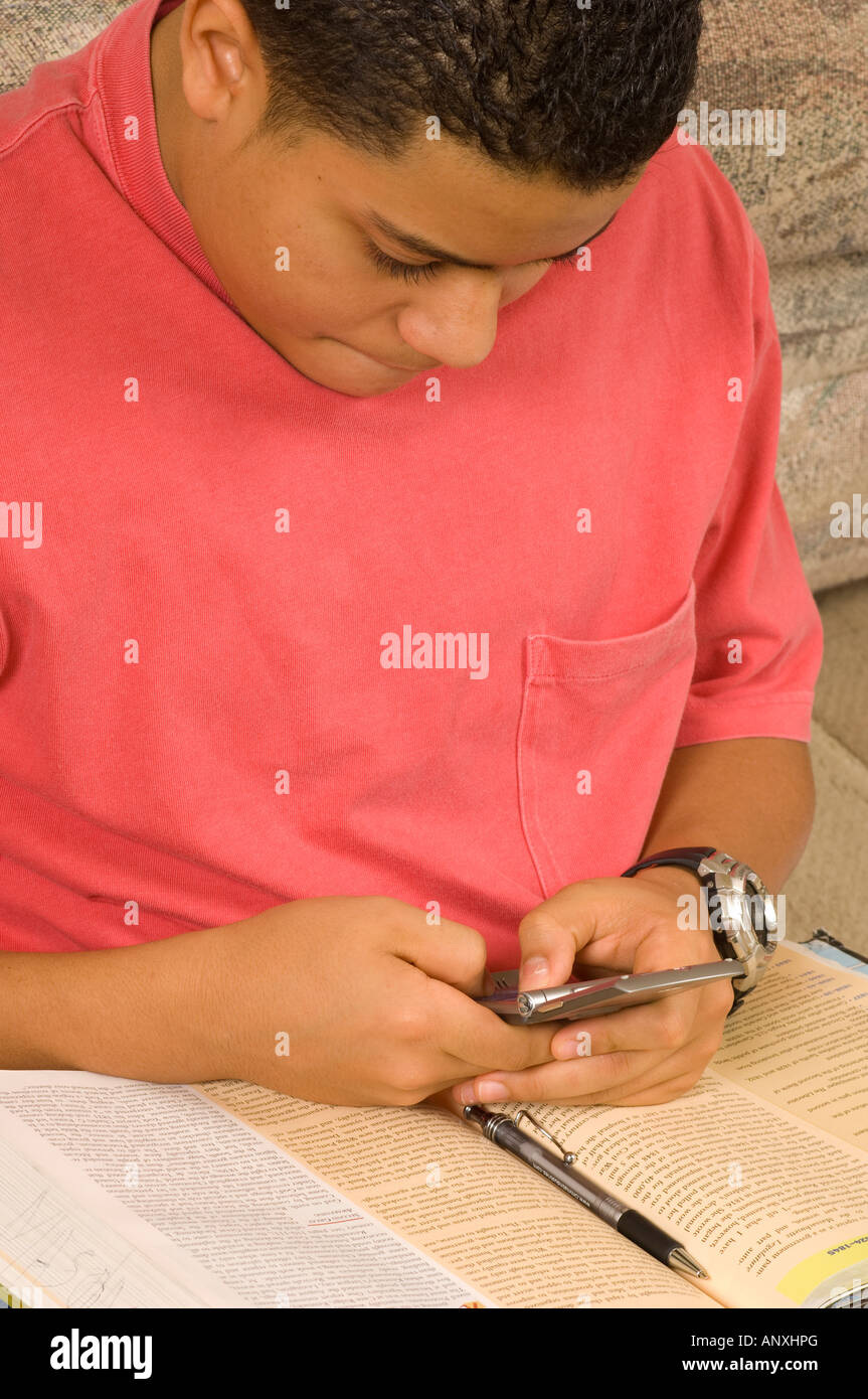 Adolescente de 15 años de edad en el hogar mediante mensaje de texto de teléfono celular deberes libro sobre lap Puertorriqueño Hispano vertical Foto de stock