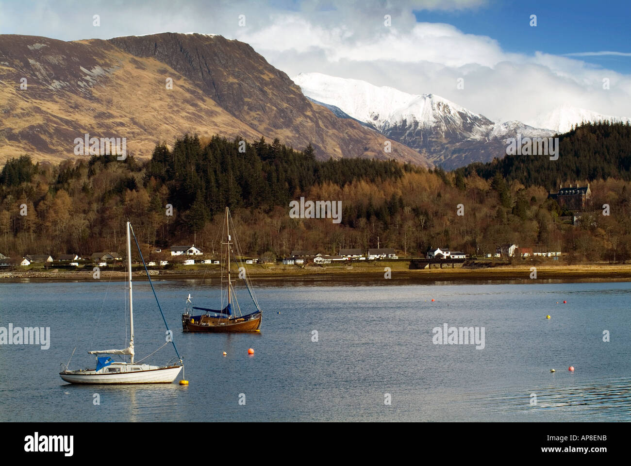 Veleros flotar suavemente en un tranquilo lago escocés Scottish dramáticos, enmarcadas por montañas nevadas y una aldea en cálidos winte Foto de stock