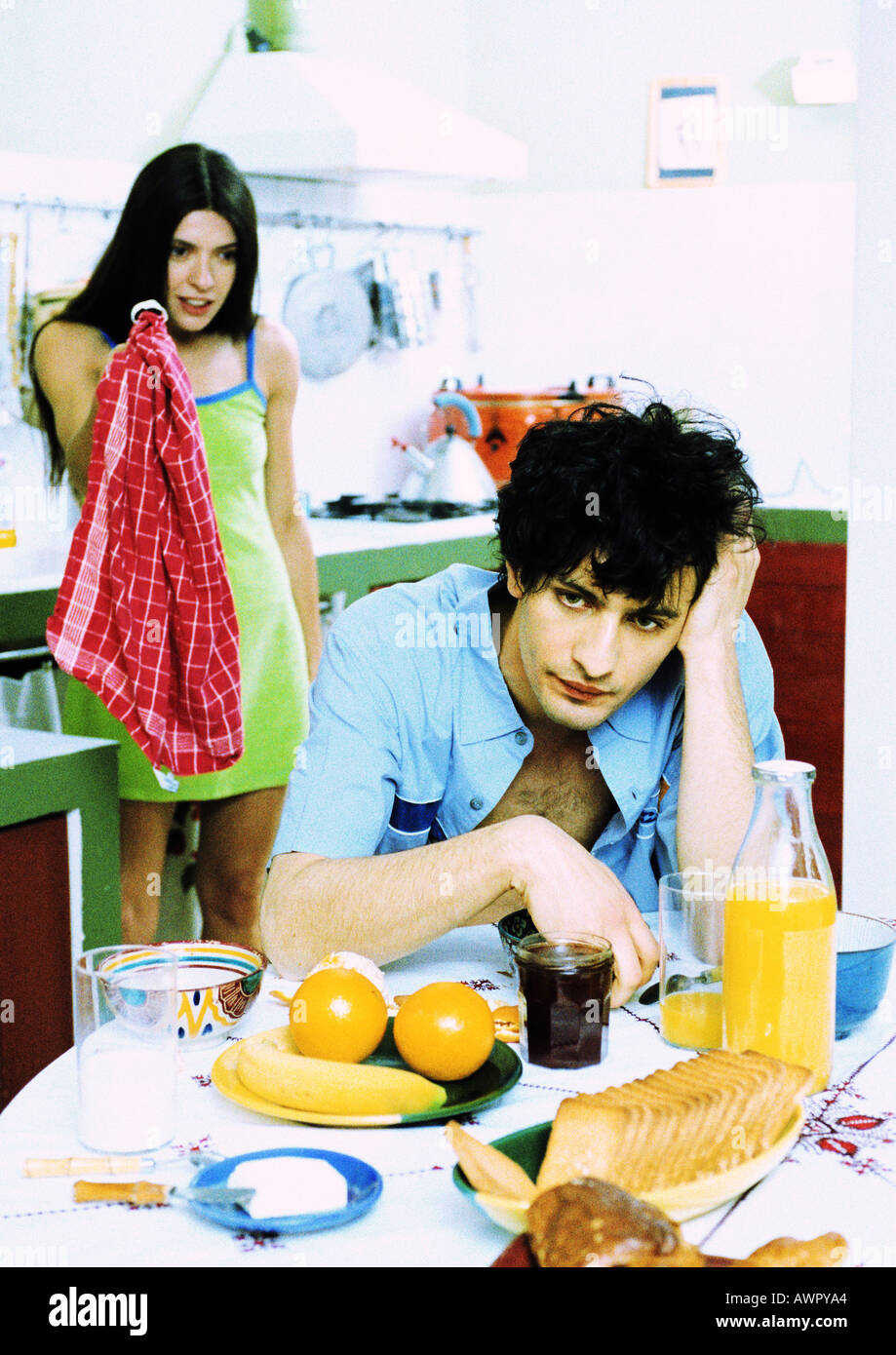 Hombre sentado en una mesa para el desayuno, mujer de pie detrás del hombre, sosteniendo una toalla de cocina. Foto de stock