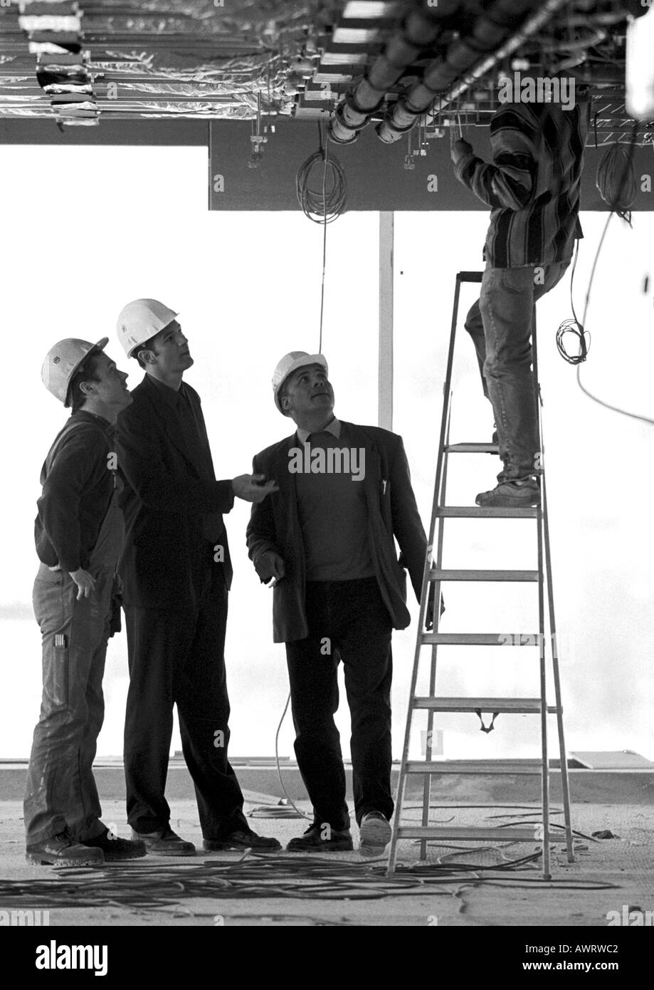 Tres hombres con sombreros duros mirando al hombre sobre la escalera, B&W Foto de stock