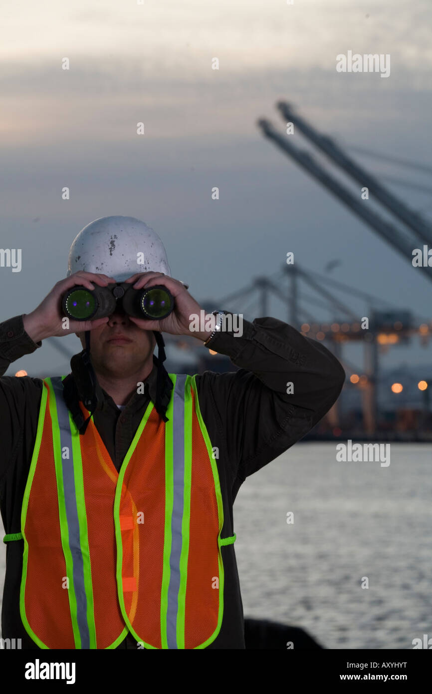 Trabajador con casco y chaleco de seguridad mirando a través de binoculares con grúas en el fondo Foto de stock