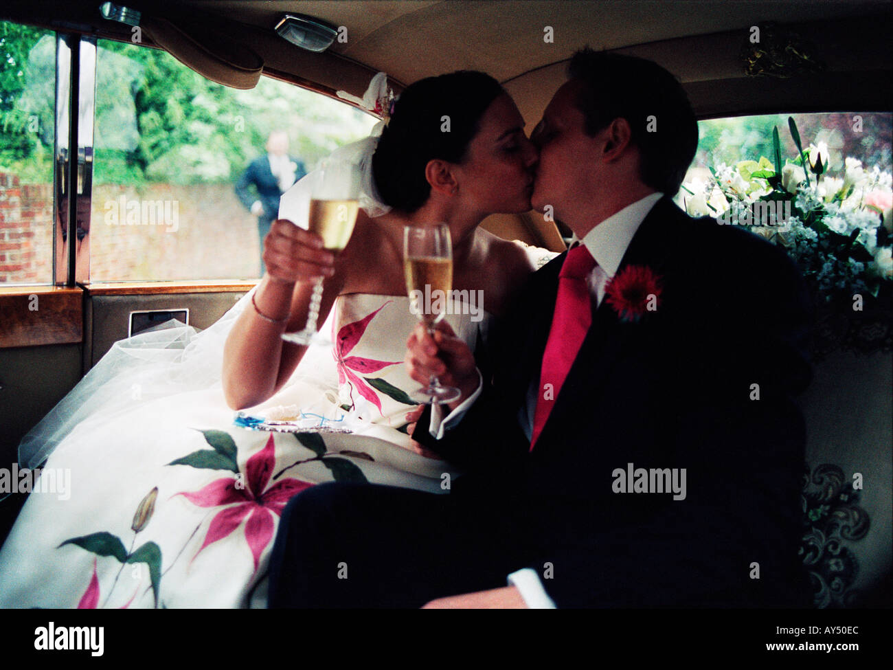 Cándido natural de la novia y el novio en la parte de atrás de su coche de boda besar mientras mantiene copas de champán Foto de stock