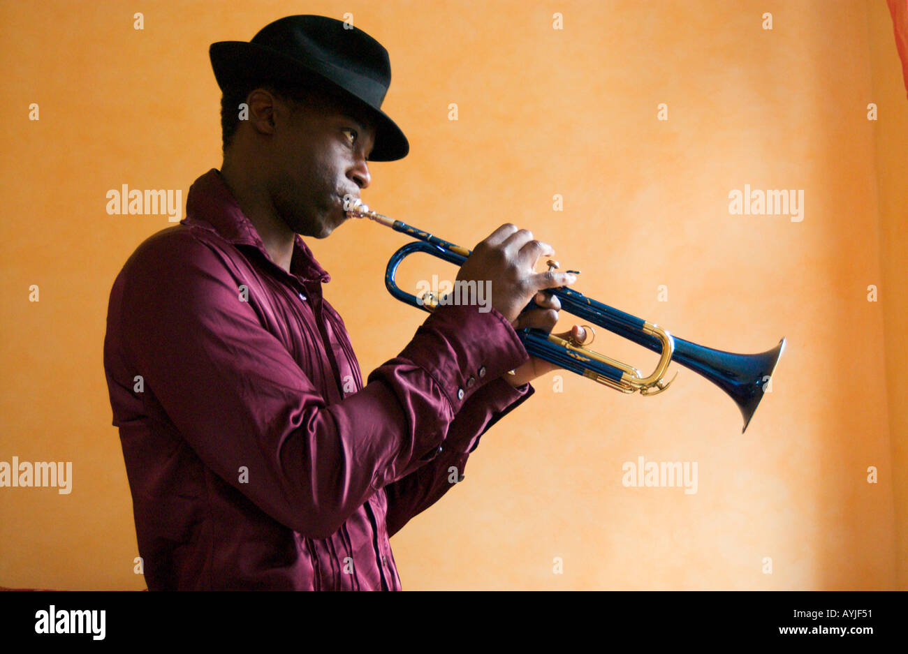 Hombre afroamericano vistiendo un sombrero Fedora desempeña una trompeta azul delante de una pared amarilla dentro Foto de stock