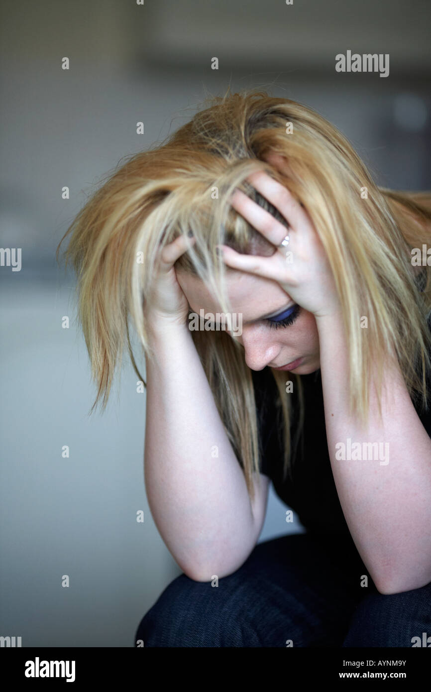 Cerca de la cabeza y los hombros de los jóvenes adolescentes de pelo rubio mujer sentada con la cabeza en sus manos, en casa, en su cocina Foto de stock