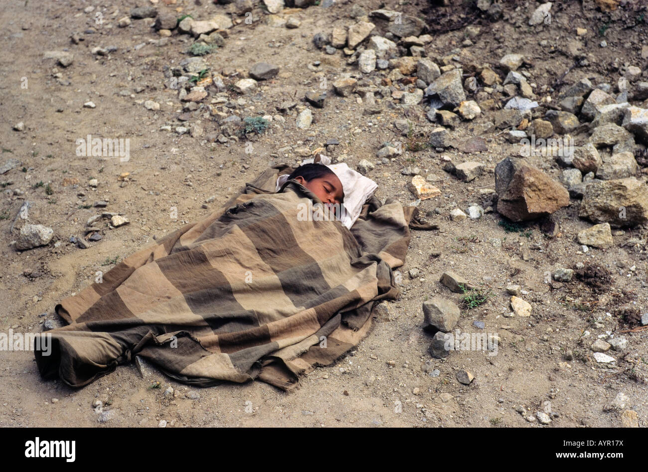 Pequeño niño envuelto en la manta de lana poniendo en un polvoriento camino de ripio para dormir, Cachemira, Himalaya, India Foto de stock