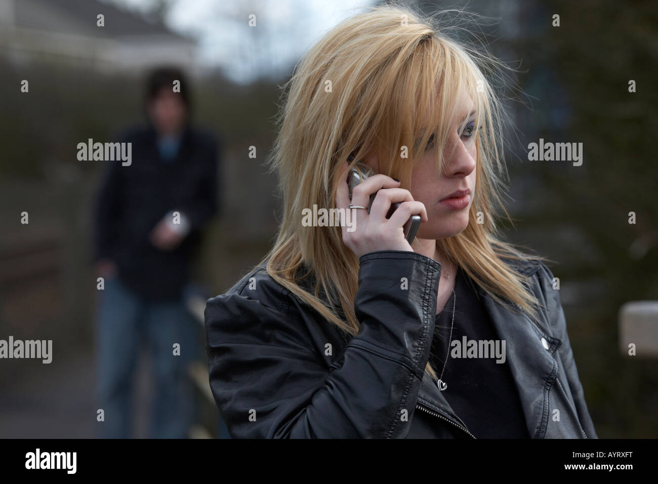 mujer adolescente rubia de pelo en el teléfono móvil haciendo una llamada de pie con el hombre de pie mirando cerca detrás de la mujer siendo acosada distanciamiento social Foto de stock