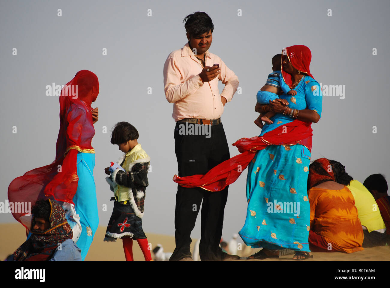 Un hombre de origen indio en ropa moderna comprueba su teléfono móvil rodeado de mujeres en prendas tradicionales en el Camel Festival en Th Foto de stock