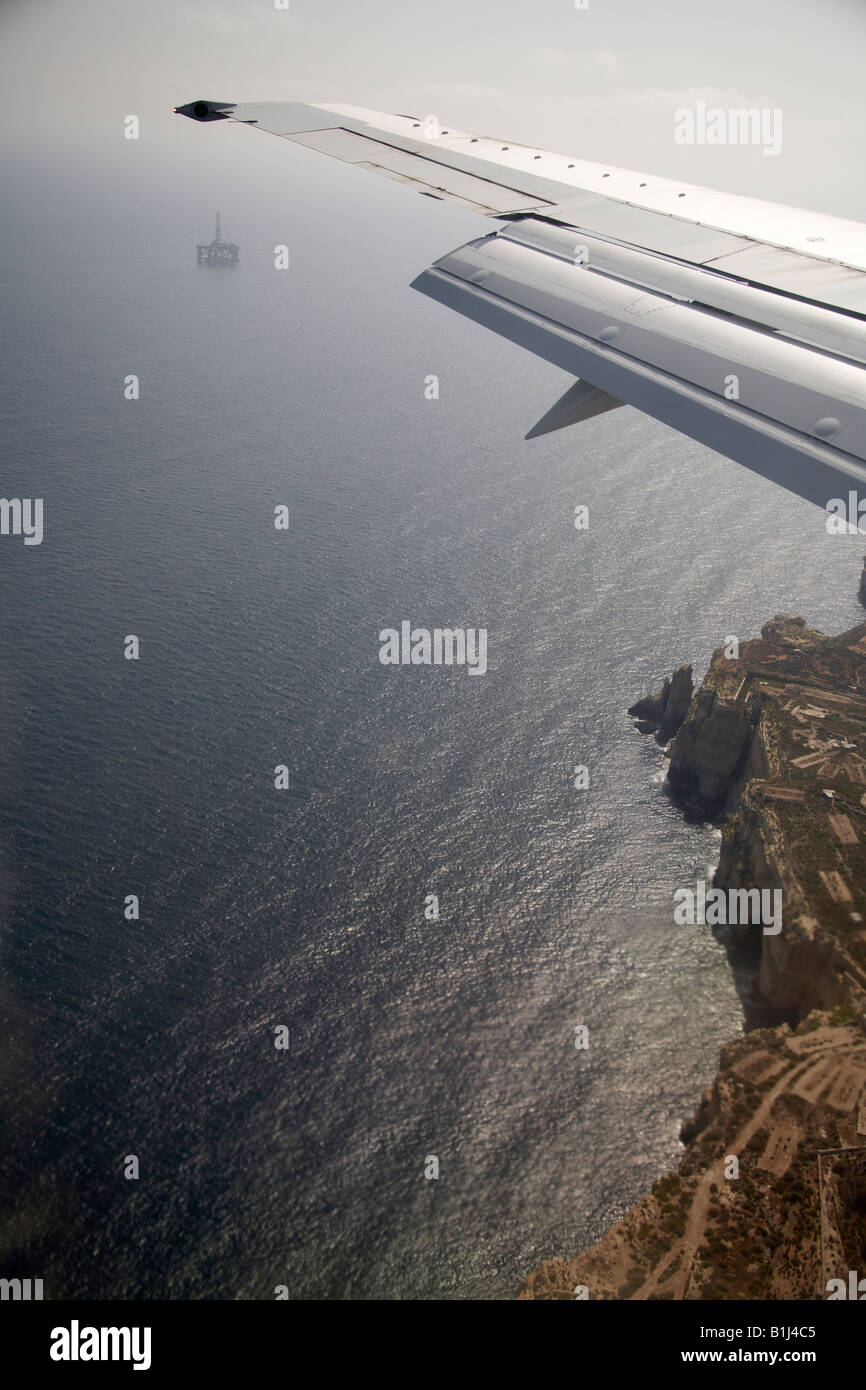 Alto nivel vista aérea oblicua de alas de avión volando sobre el mar y acantilados costeros de Malta con la plataforma petrolífera en el mar Foto de stock