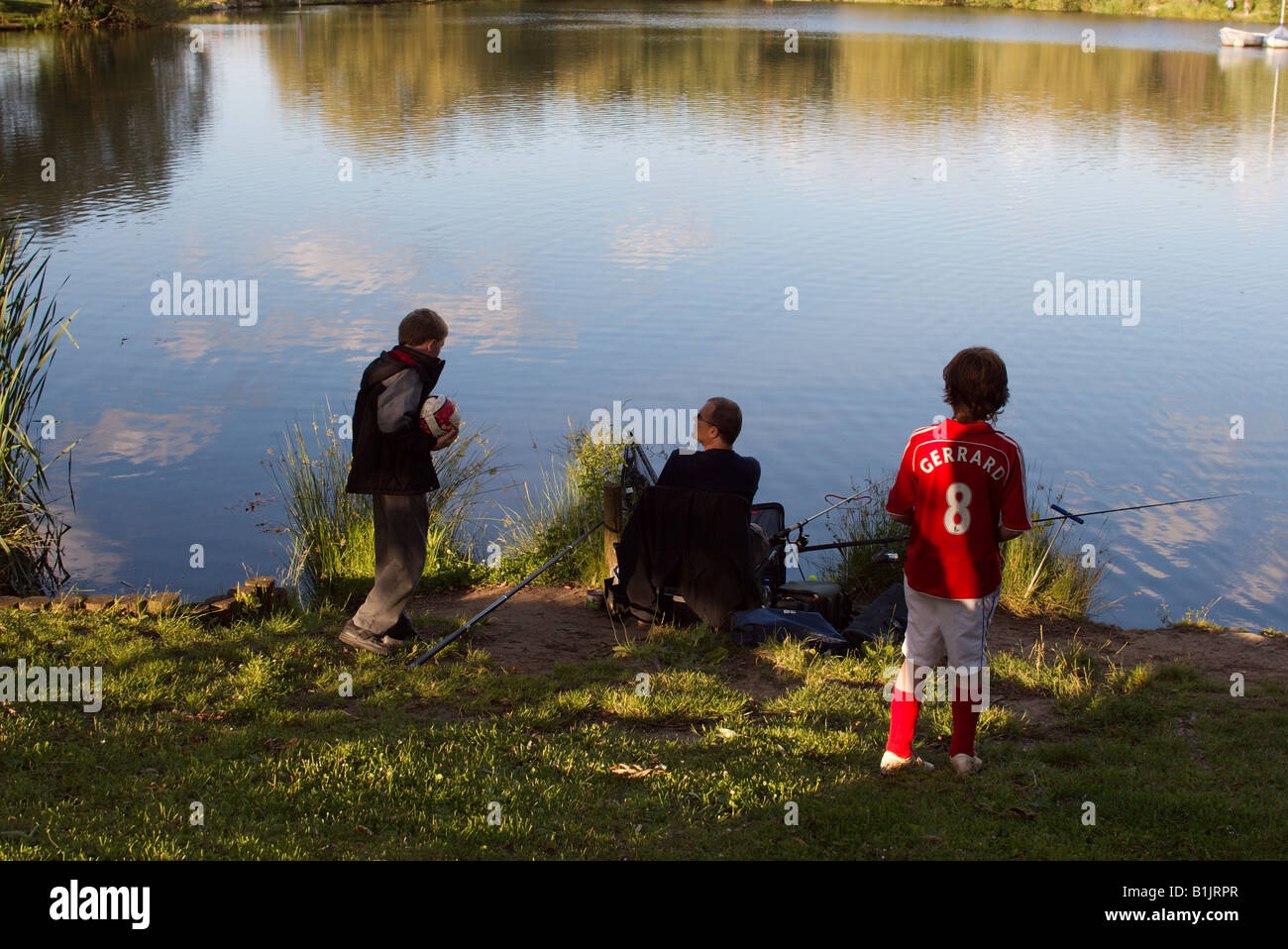 De Woking (Surrey, Inglaterra Goldsworth Park Lake pescador y dos muchachos curiosos uno busca en la red mantenga uno en una camiseta de fútbol Foto de stock