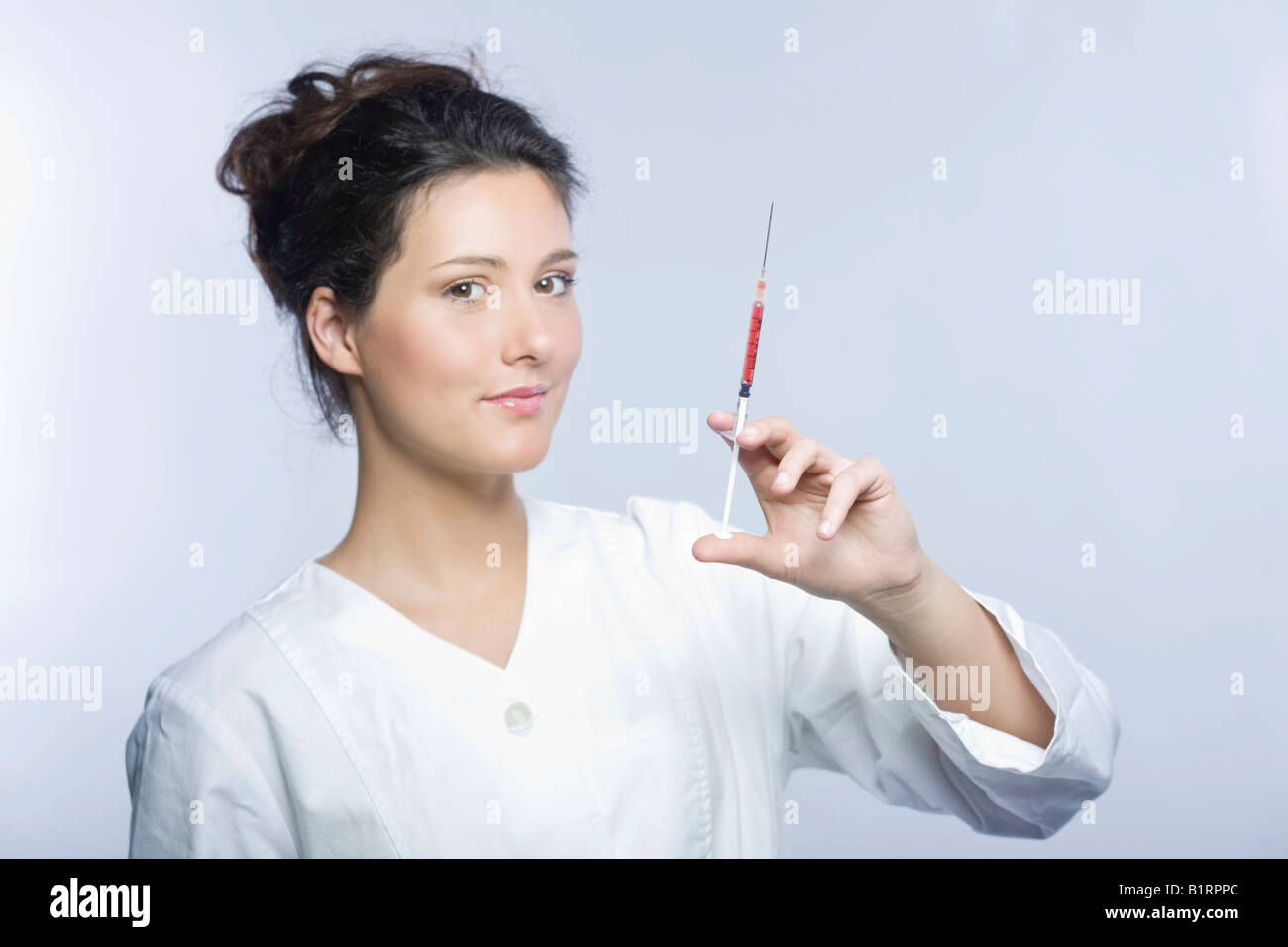 Mujer joven vestida de blanco smock laboratorio sosteniendo una jeringa llena de líquido rojo en su mano Foto de stock