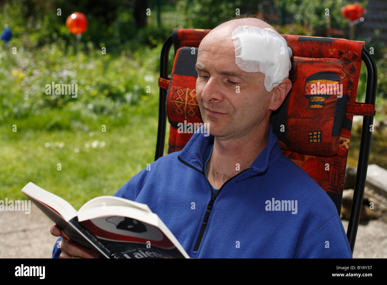 El hombre, de 45 años, con una venda adhesiva sobre su cabeza, Sentado en una silla de jardín leyendo un libro, Geretsried, Baviera, Alemania, Europa Foto de stock