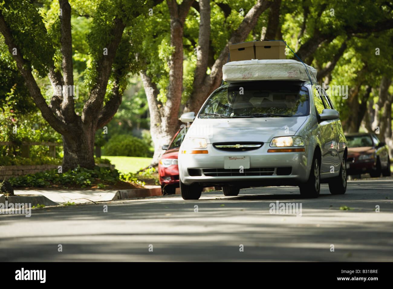 Chevy Aveo bajando la calle arbolada con colchón y cajas en el techo Foto de stock