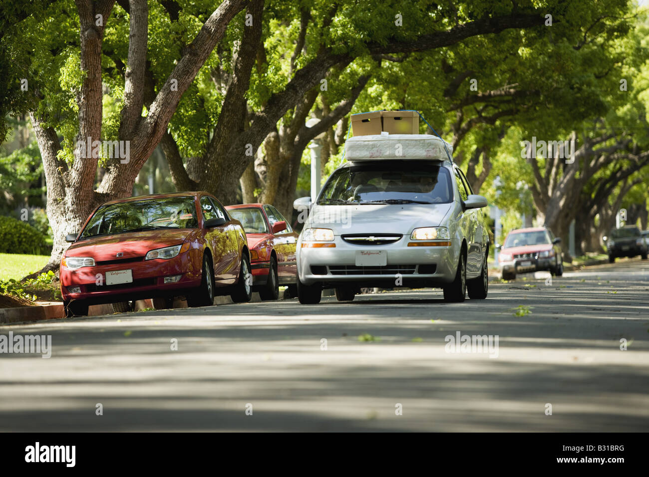 Chevy Aveo bajando la calle arbolada con colchón y cajas en el techo Foto de stock