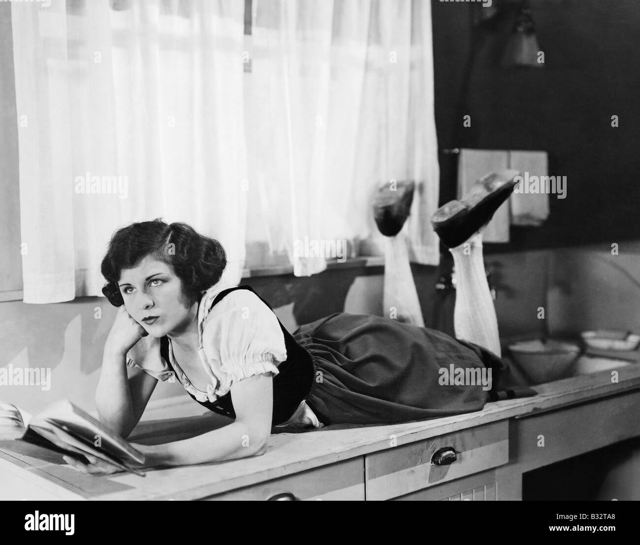 Mujer joven tumbado sobre cocina sosteniendo el pensamiento del libro Foto de stock