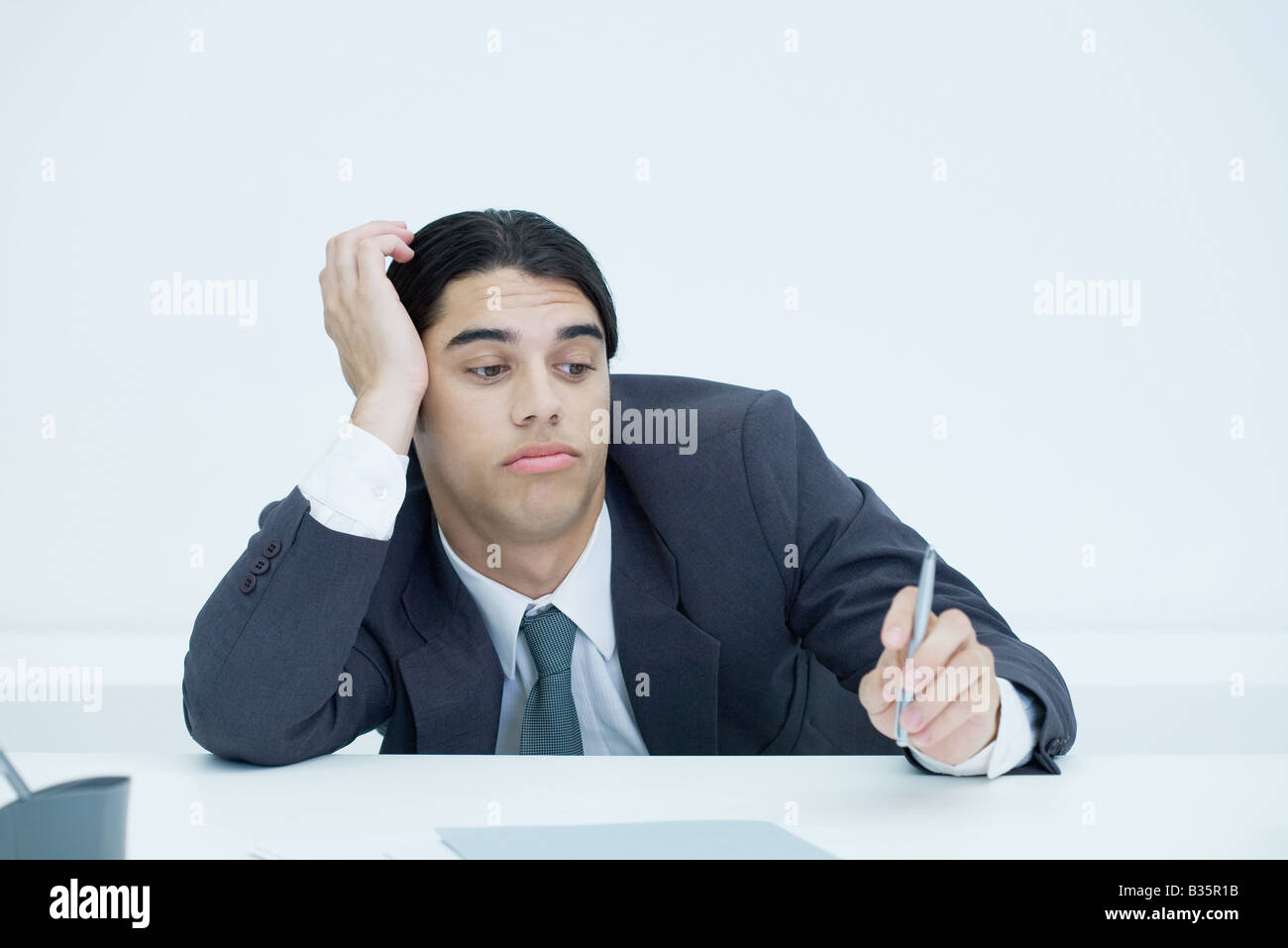 Joven Empresario de encorvamiento al escritorio, sosteniendo la cabeza y mirando con expresión pen aburrido Foto de stock