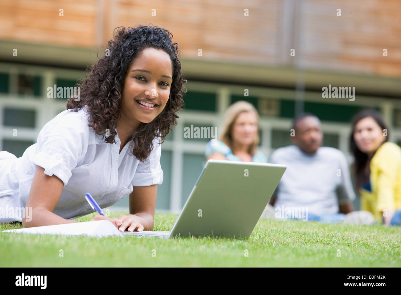 Estudiante al aire libre sobre césped utilizando portátil con otros estudiantes en el fondo (enfoque selectivo) Foto de stock
