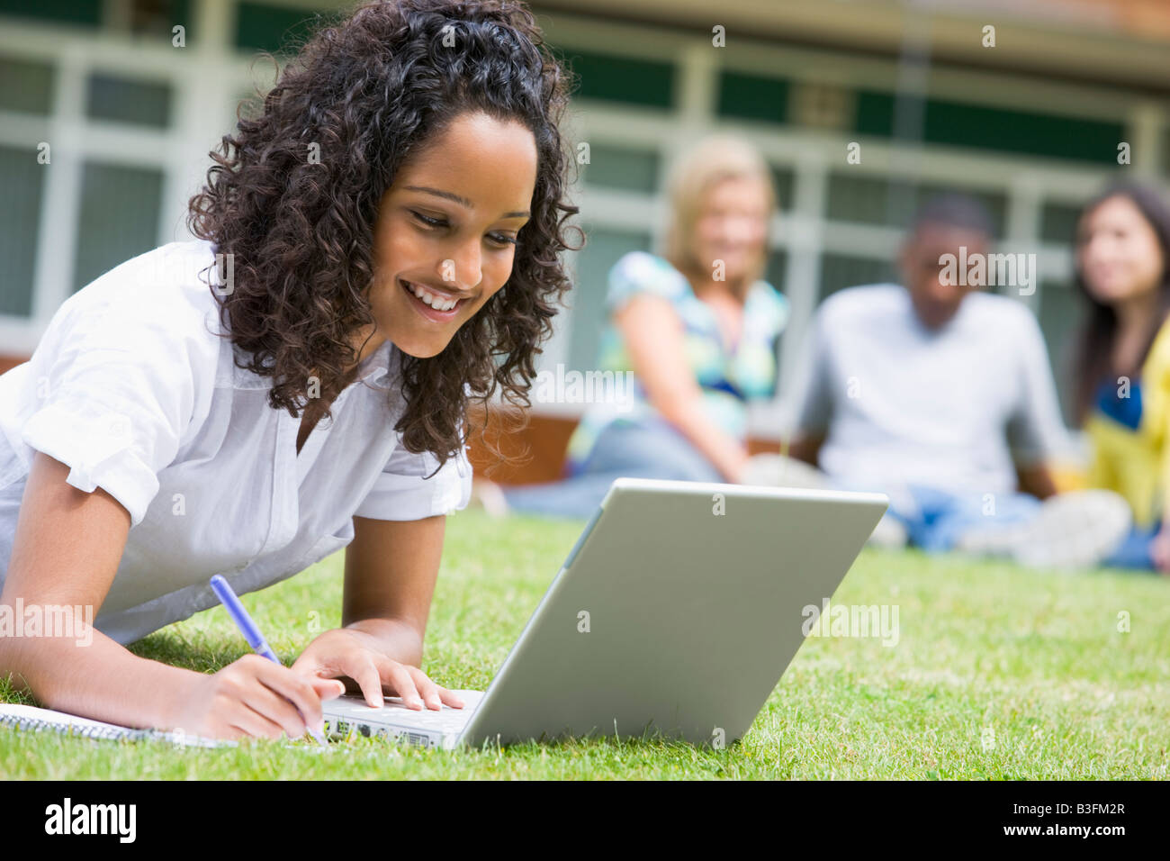 Estudiante al aire libre sobre césped utilizando portátil con otros estudiantes en el fondo (enfoque selectivo) Foto de stock