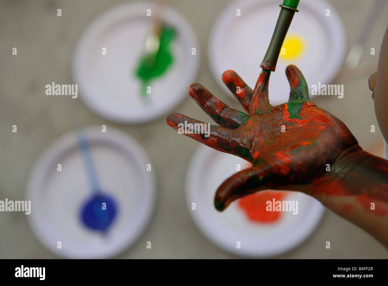 Un niño preescolar experimentos por pintar su mano con diferentes colores primarios, haciendo una paleta desordenada. Foto de stock