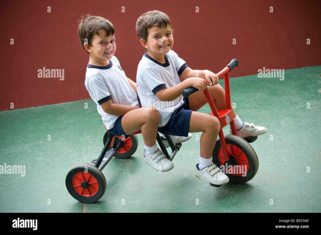 GEMELOS NIÑOS IDÉNTICOS 4-6 años Feliz sonrisa gemelos idénticos, niños pequeños jugando en bicicleta para dos fuera en el patio de recreo de la escuela jardín de infancia Foto de stock
