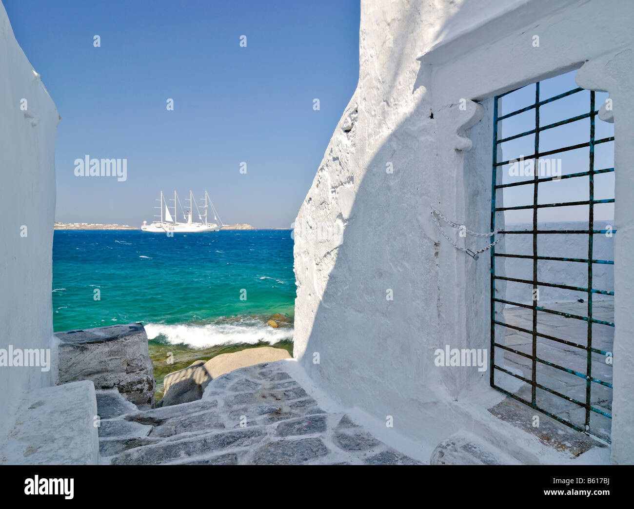 Vista a través de un callejón con paredes blancas y una puerta con barras hacia un velero de cuatro mástiles o barca en un mar turquesa Foto de stock