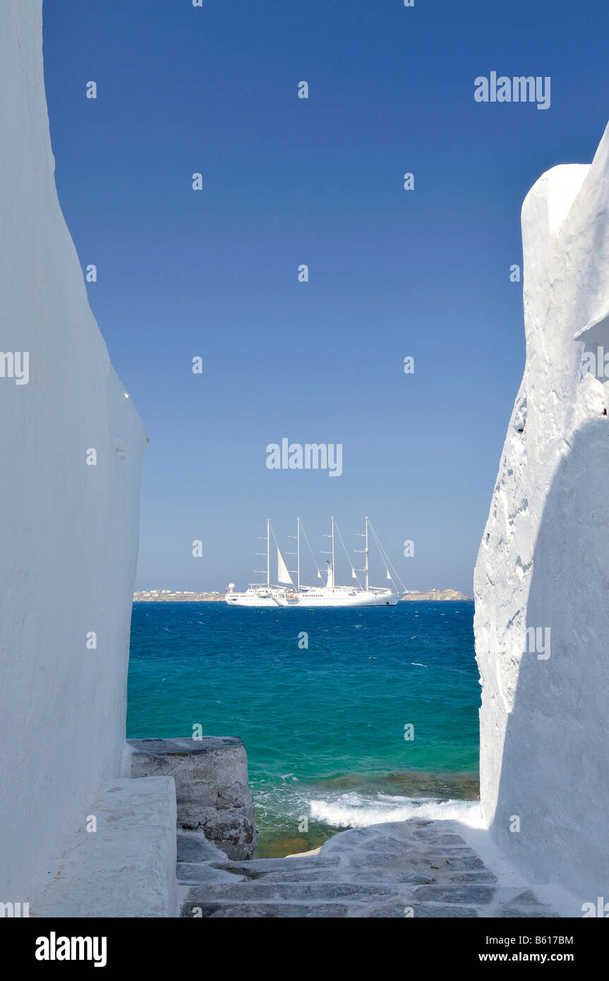 Vista a través de un callejón con paredes blancas hacia un velero de cuatro mástiles o barca en un mar azul turquesa, Mykonos Cyclades Foto de stock