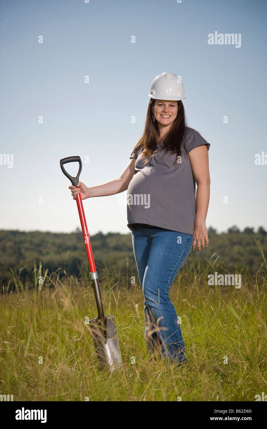 Joven Embarazada de pie en un campo con una pala y vistiendo un sombrero duro. Foto de stock