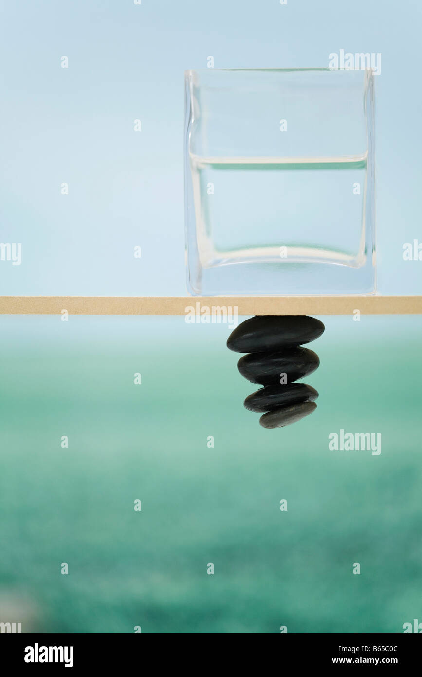 Recipiente de vidrio lleno con agua sentado en la cornisa, el montón de piedras desafiando la gravedad debajo Foto de stock
