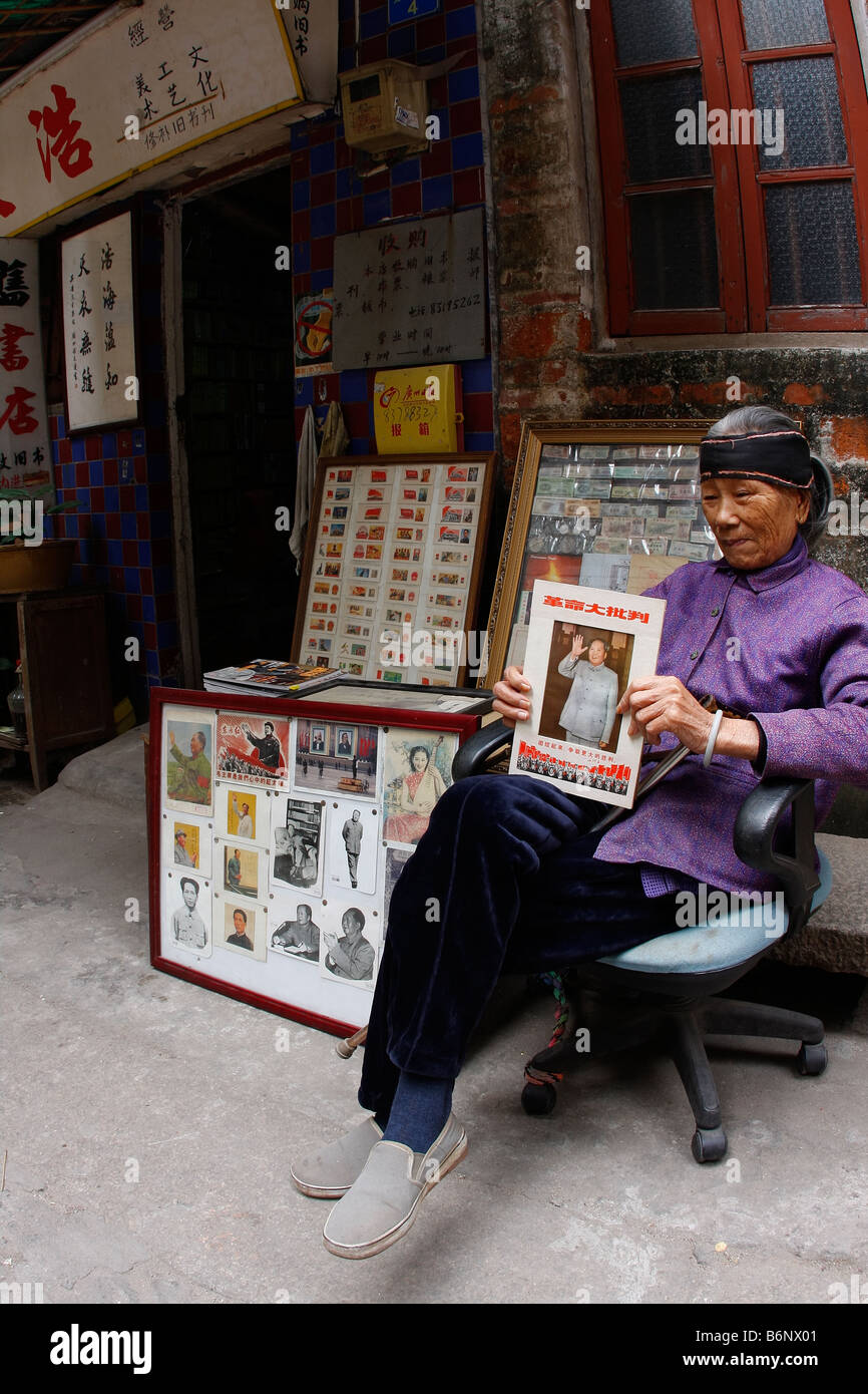 Un muy lindo hermoso y elegante dama en chino de 93 años sentada enfrente de su tienda en Guangzhou con propaganda de Mao. Foto de stock