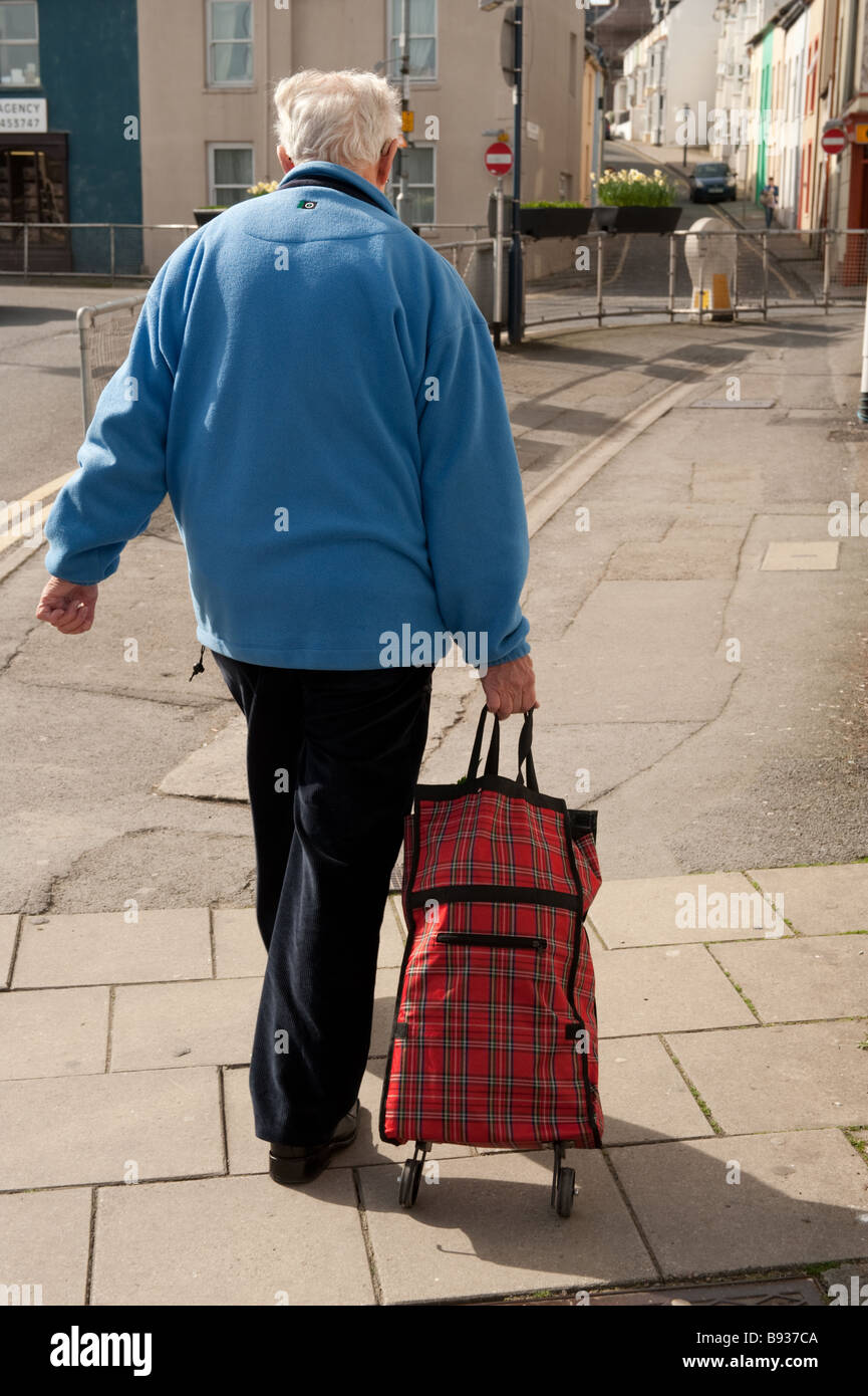 Vista trasera desde detrás de un anciano caminando a casa con sus compras en un tartan bolsa con ruedas Foto de stock